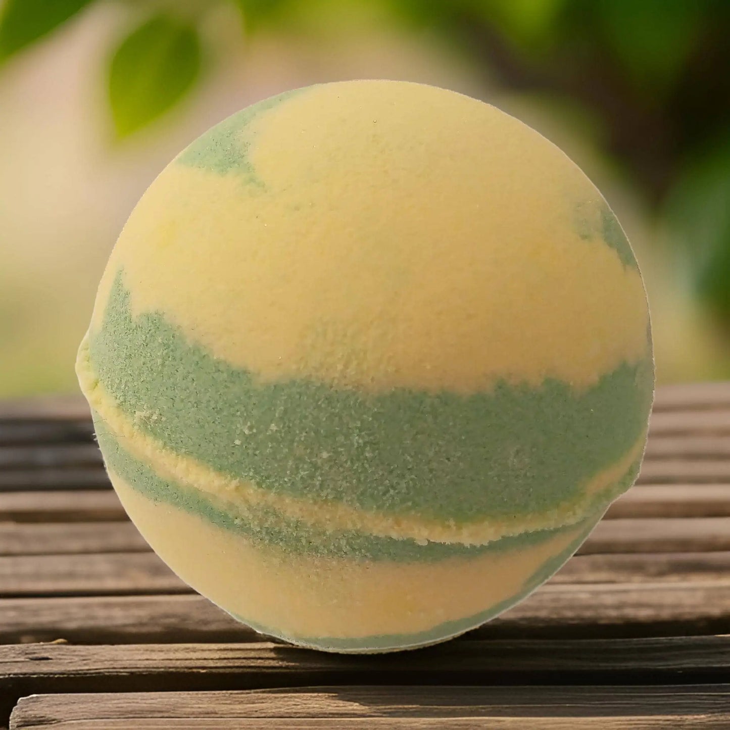A round Gooseberry & Elderflower Fizzy Bath Bomb by Posh Brats, featuring yellow and green swirls, sits on a wooden surface against a blurred natural backdrop. Ideal for aromatherapy, it turns your bath into a refreshing getaway.