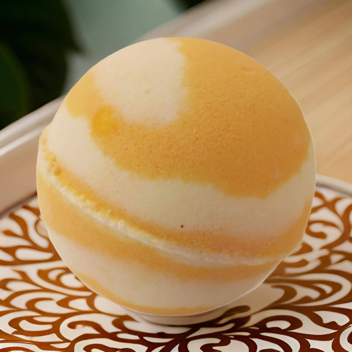 A round, orange and cream Coconut & Pineapple Fizzy Bath Bomb by Posh Brats sits on a decorative tray with swirling brown patterns. Resembling a planet with smooth gradients and a visible seam, it offers a luxurious bath experience infused with natural oils and botanical extracts.