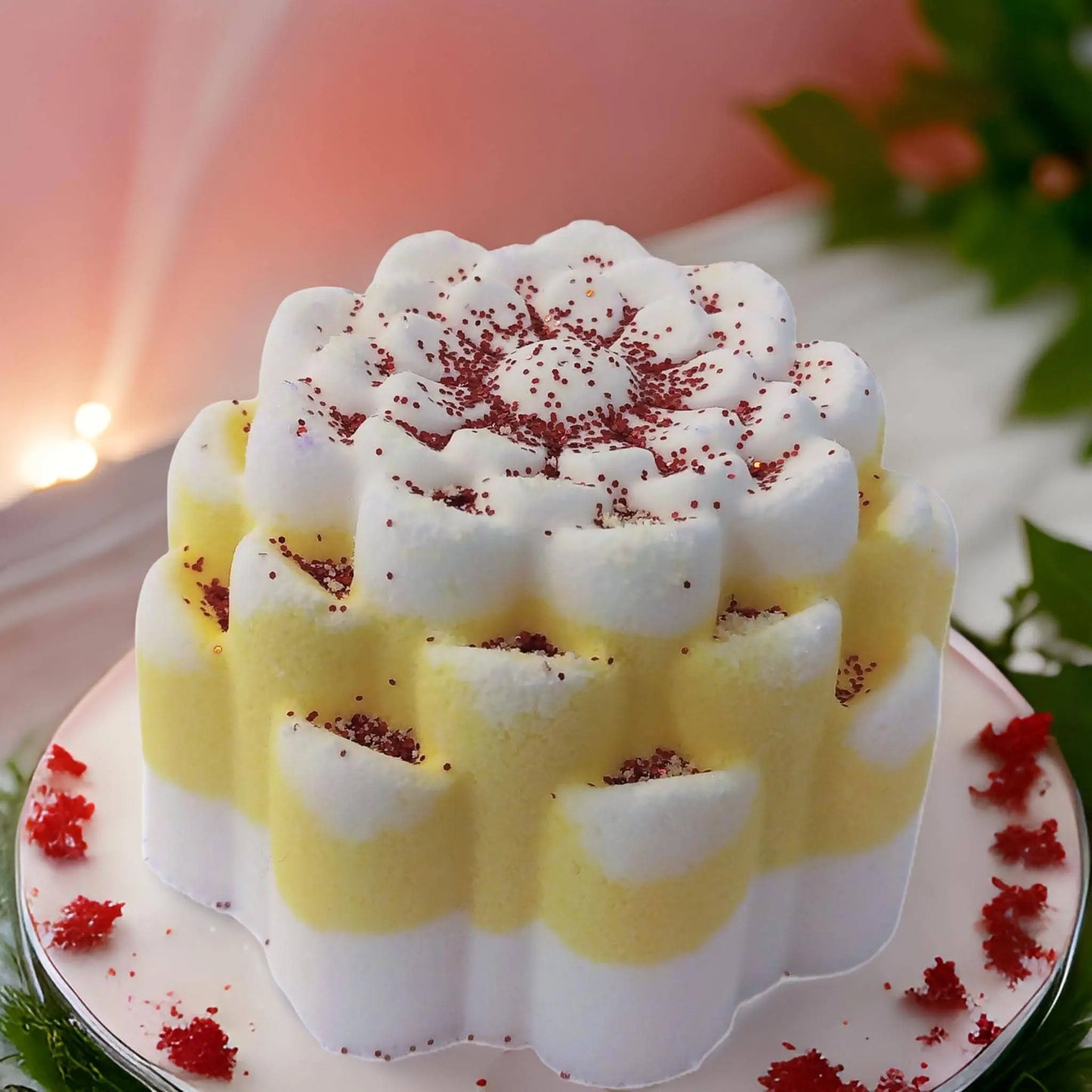 The Pina Colada Fizzy Bath Bomb by Posh Brats rests beautifully with its decorative flower shape and white-yellow layers, topped with red sprinkles. Surrounded by green leaves and red petals, it sits on a white plate against a pink, softly lit backdrop.