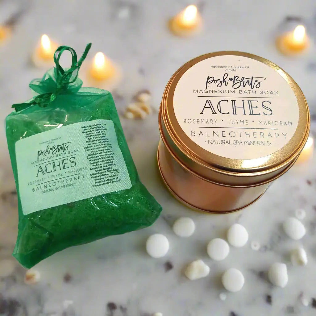 A green bag of Posh Brats Aches Aromatherapy Bath Salt and a gold tin, infused with rosemary, thyme, marjoram, and eucalyptus, rest on marble. Soft candlelight and white stones create an elegant spa-like ambiance.