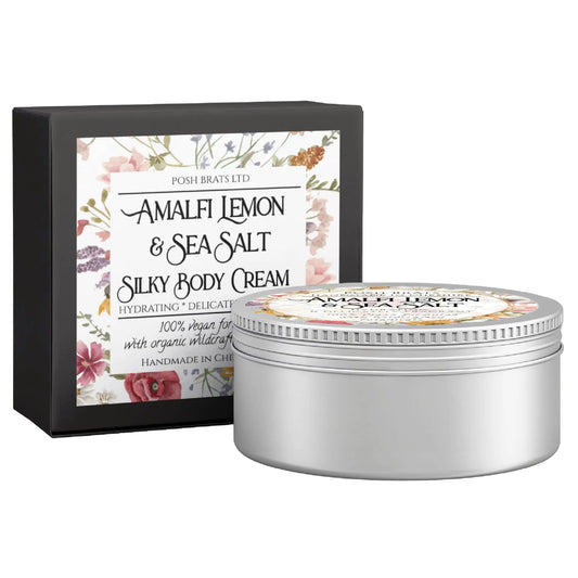 A jar of Posh Brats Amalfi Lemon + Sea Salt Silky Body Butter Cream sits elegantly before its decorative box, which features floral artwork and product details, highlighting the hydrating formula crafted with organic wildcrafted oils.