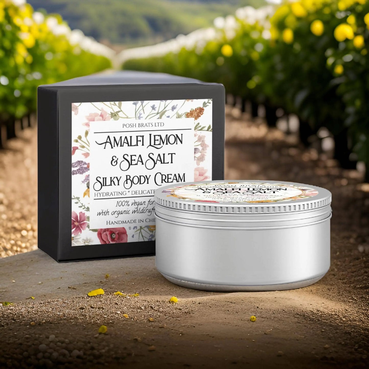 A jar and box of Amalfi Lemon + Sea Salt Silky Body Butter Cream from Posh Brats sit on a dirt path, with a lush, green field dotted with vibrant yellow flowers stretching under a clear sky in the background.