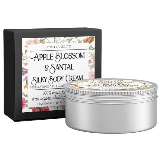 The image shows a round tin of Apple Blossom + Santal Silky Body Butter Cream by Posh Brats next to its black box. The box, decorated with a floral design, highlights the products hydrating qualities and its vegan, organic ingredients.