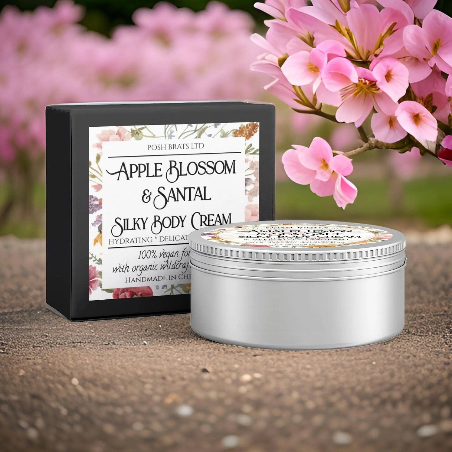 A decorative tin of Apple Blossom + Santal Silky Body Butter Cream by Posh Brats sits before its matching box, set against blooming pink flowers and a blurred natural landscape, promising hydrating skin nourishment.