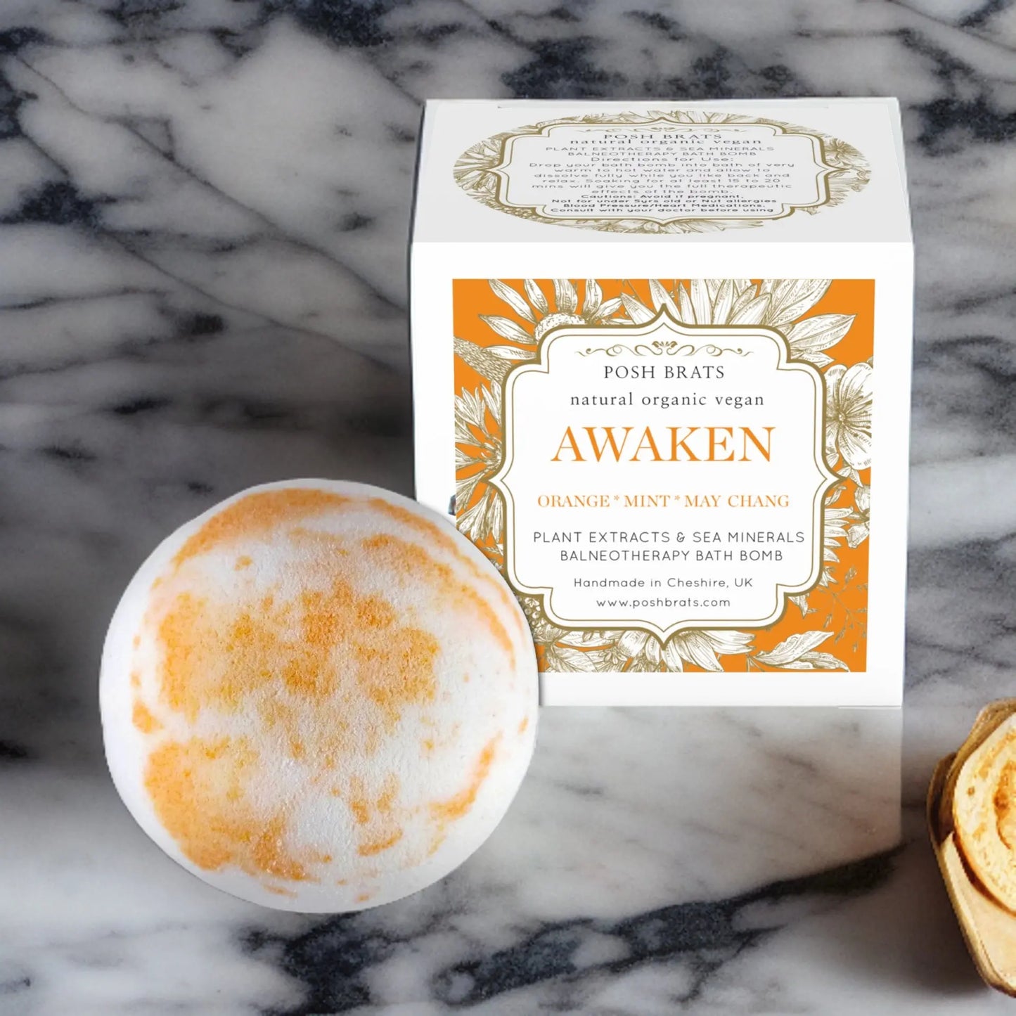The Awaken Aromatherapy Bath Bomb from Posh Brats, round and orange-white, rests near a box listing its essential oils—orange, mint, may chang. Touted as a natural vegan product, it promises a rejuvenating bath experience on marble.