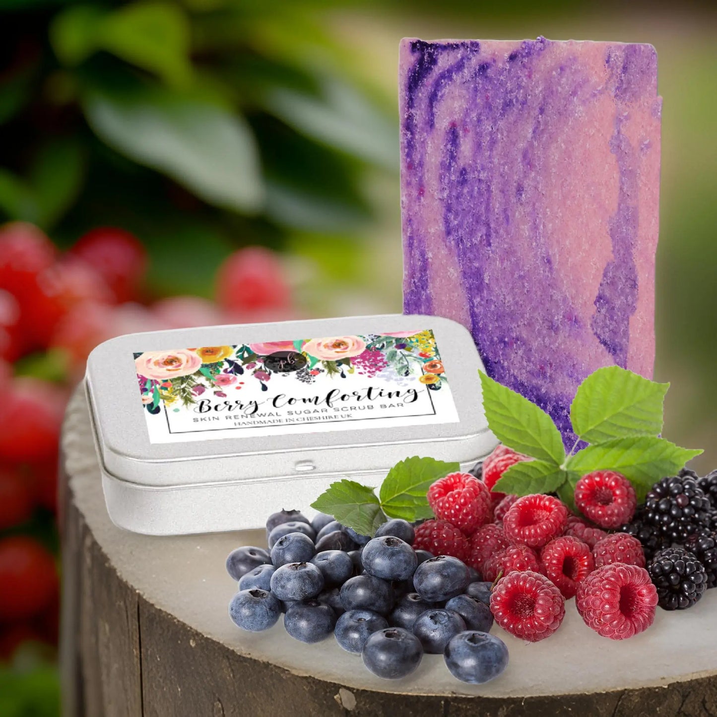 A Berry Comforting Sugar Scrub Bar from Posh Brats, with a purple and pink swirl, rests on a wooden surface adorned with blueberries, raspberries, and blackberries. Beside the Berry Comforting tin, lush greenery enhances the natural feel of this skincare scene.