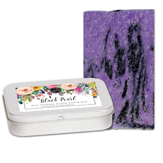 A purple sugar scrub bar with black swirls stands upright next to a rectangular tin with a floral label reading Black Pearl Skin Renewal Sugar Scrub Bar. Made by Posh Brats, it features organic British beet sugar and vegan exfoliating properties, while the container has a sleek silver finish.