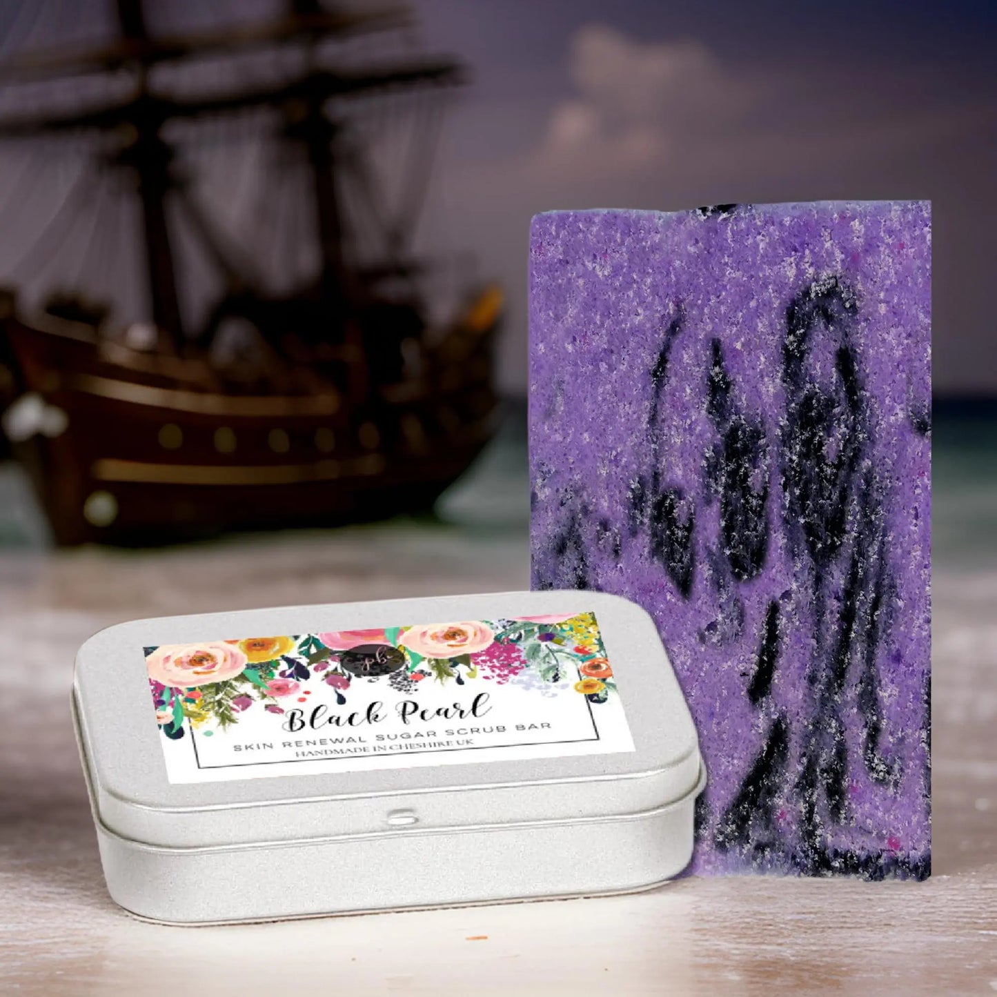 A purple soap bar with black swirls sits beside a silver tin on sand. The label reads Black Pearl Sugar Scrub Bar by Posh Brats, made with organic British Beet Sugar. A blurred wooden ship model in the background adds nautical charm.