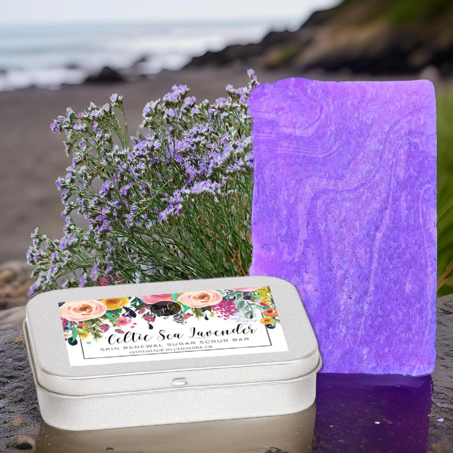 A bright purple sugar scrub bar made with organic sugar crystals rests next to a silver tin labeled Celtic Sea Lavender Sugar Scrub Bar by Posh Brats. The peaceful scene features a rocky beach with delicate purple flowers.