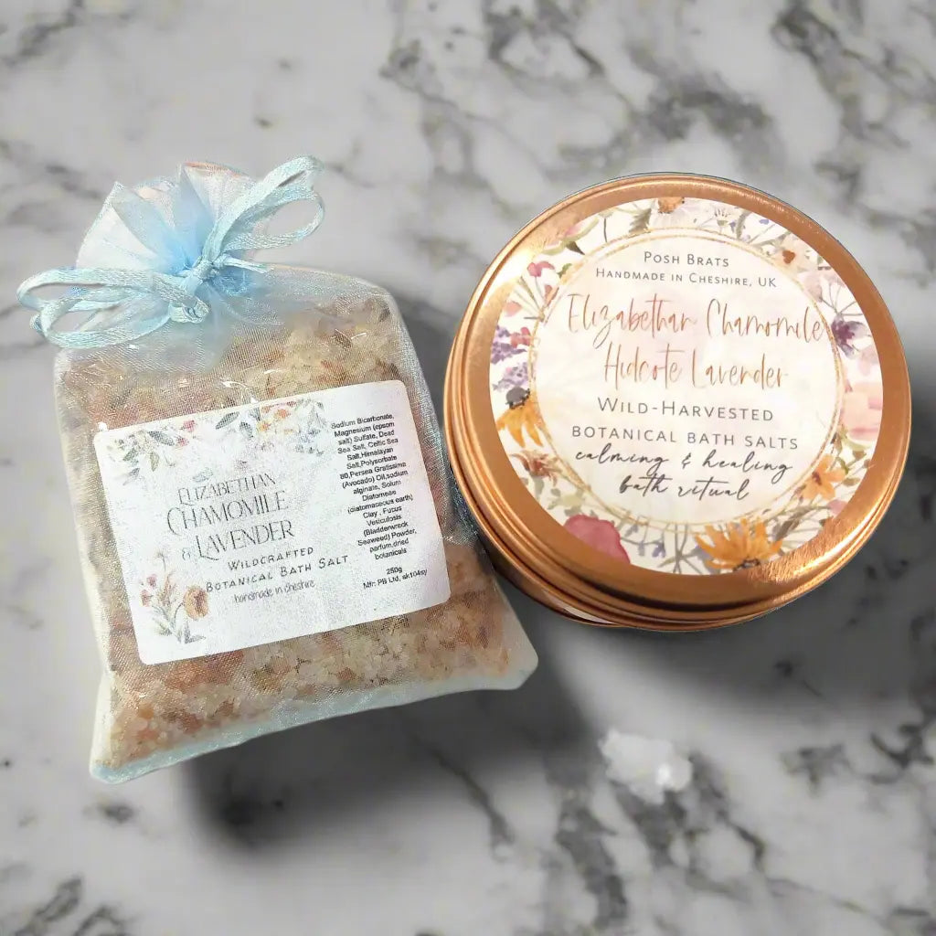 A sheer blue pouch of Elizabethan Chamomile Lavender Bath Salt from Posh Brats sits beside a round floral tin. Labeled Wild-Harvested Botanical Bath Salts, it promises the same soothing scent for an indulgent bathing experience.