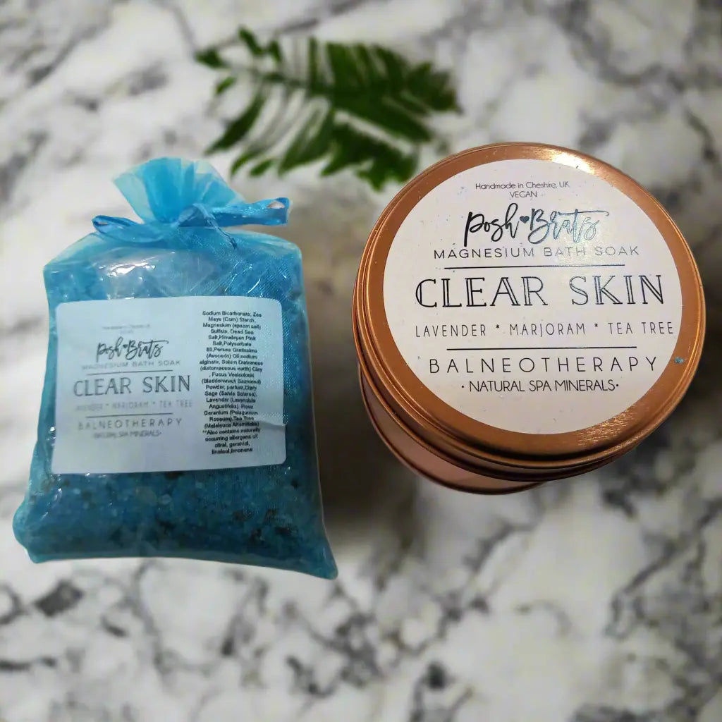 On a marble surface sits a blue bag of Posh Brats Clear Skin Magnesium Aromatherapy Bath Salt with lavender, marjoram, and tea tree. Nearby, round containers labeled the same are stacked with a green leaf for a natural touch in the background.