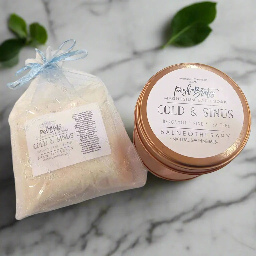 A marble surface showcases Posh Brats Cold Sinus Magnesium Bath Salt in a fabric pouch and tin container. The pouch contains bath salts, while the tin offers a Cold Sinus soak infused with eucalyptus and peppermint oils, with green leaves artfully arranged in the background.