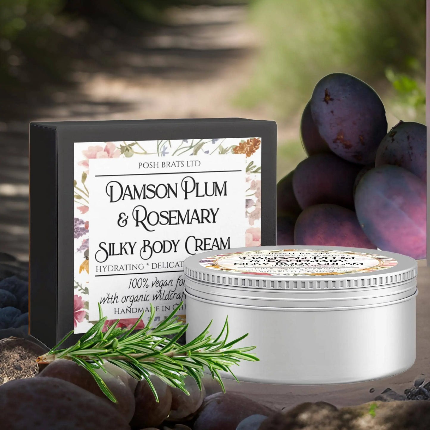 A tub and box of Posh Brats Damson Plum + Rosemary Silky Body Butter Cream sit on a dirt path surrounded by fresh plums and rosemary. This vegan-friendly, hydrating cream is crafted with organic ingredients.