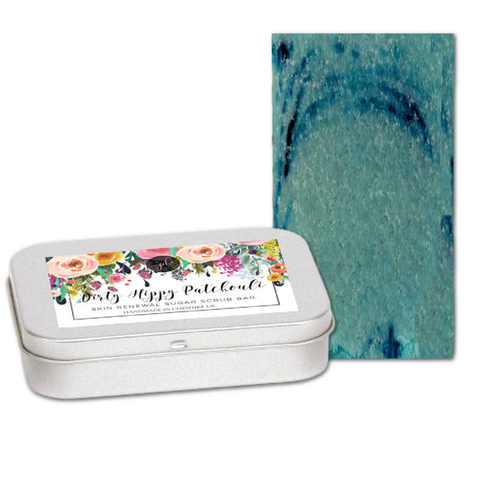 A rectangular metal tin with a floral label reads Dirty Hippy Patchouli Sugar Scrub Bar by Posh Brats, offering a delightful skin renewal. A textured teal-blue bar promises delicately exfoliated skin with every use, enriched with invigorating patchouli essence.