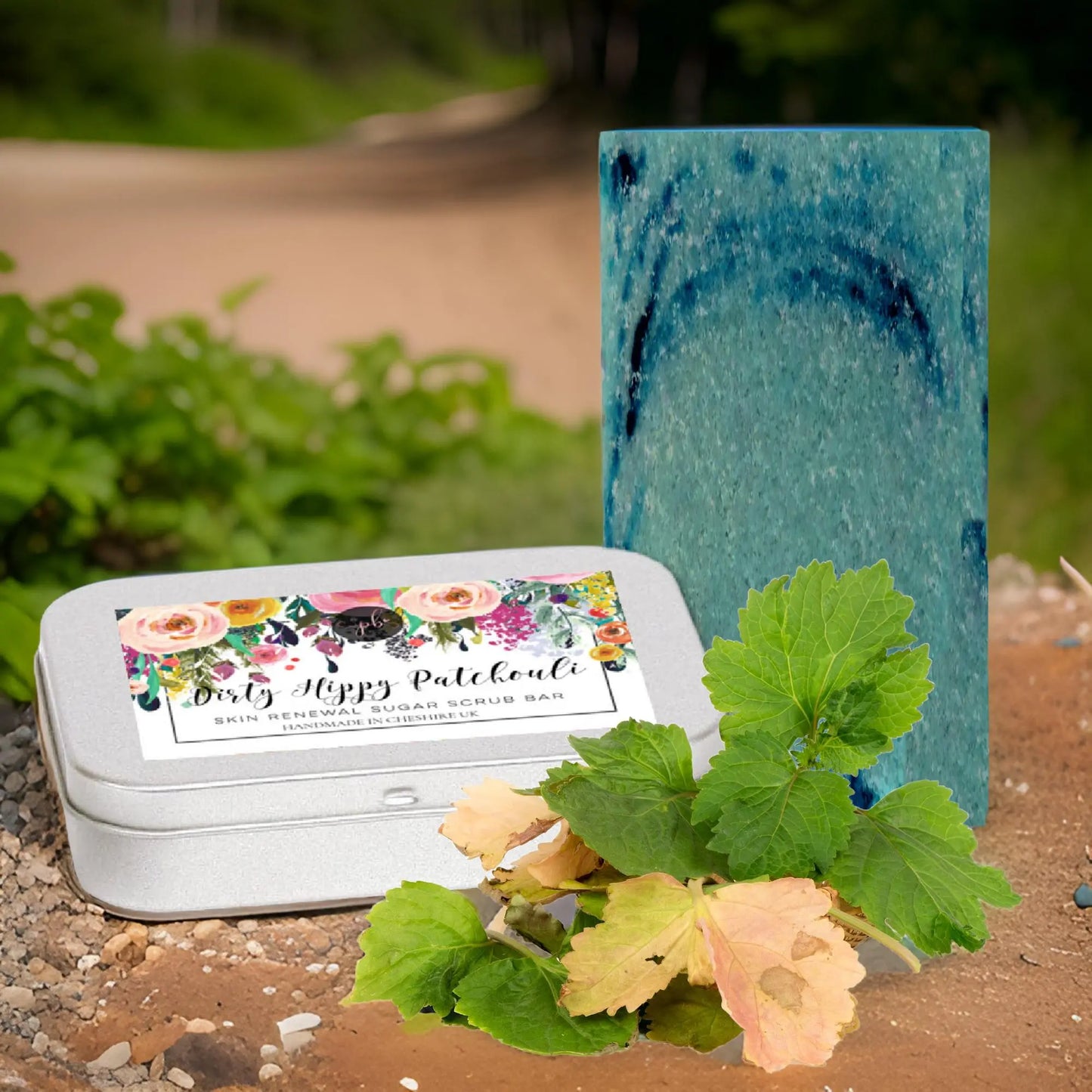 A Dirty Hippy Patchouli Sugar Scrub Bar by Posh Brats sits in a metal tin on a dirt path surrounded by lush foliage. Nearby, a blue soap bar stands tall against trees, promising to exfoliate and invigorate your skin with earthy patchouli notes.