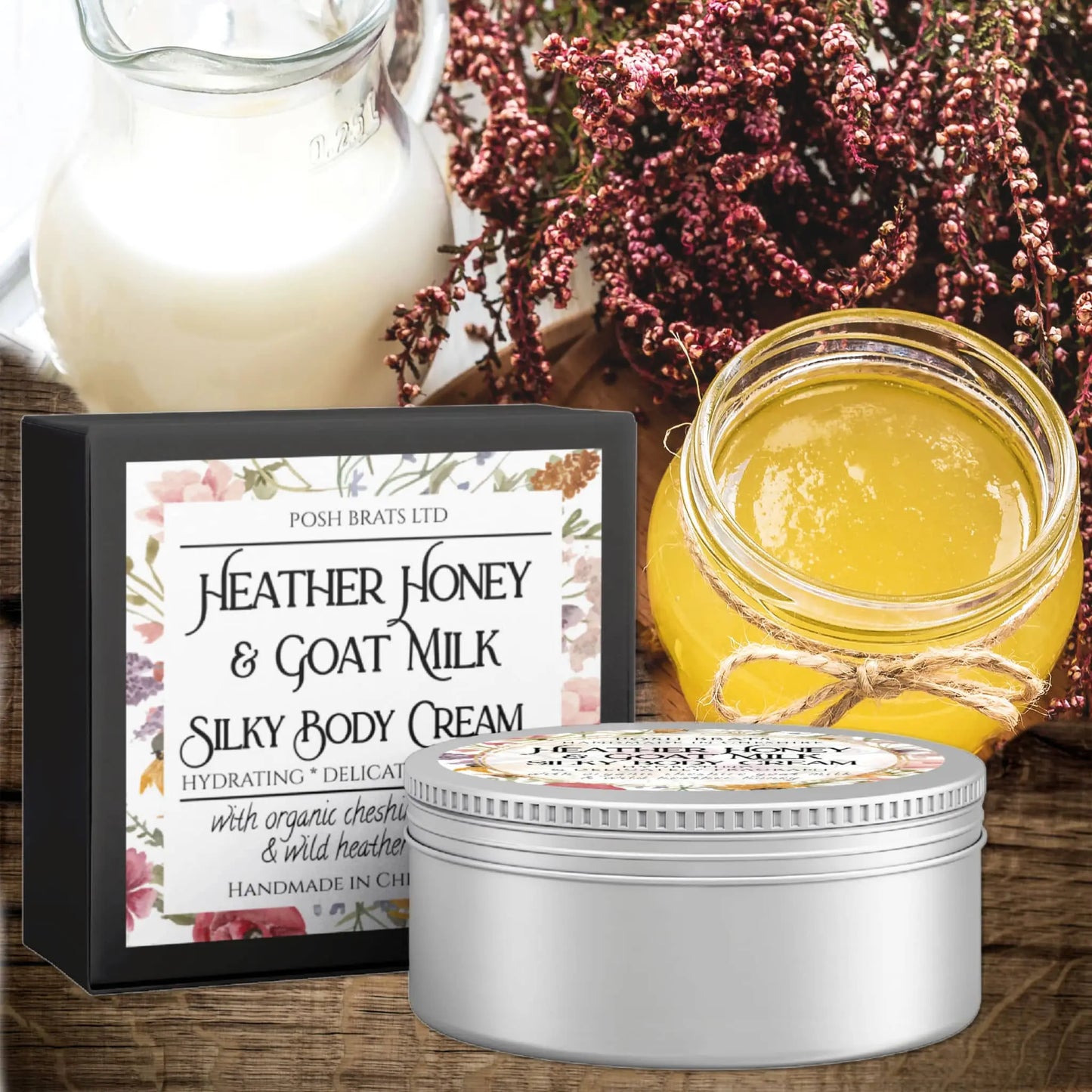 The Heather Honey + Goat Milk Silky Body Butter Cream by Posh Brats features elegant packaging. Nearby, a jar of golden honey, rich milk, and wild heather completes the scene. This hydrating cream is crafted with organic chestnut honey and delicate wild heather notes.