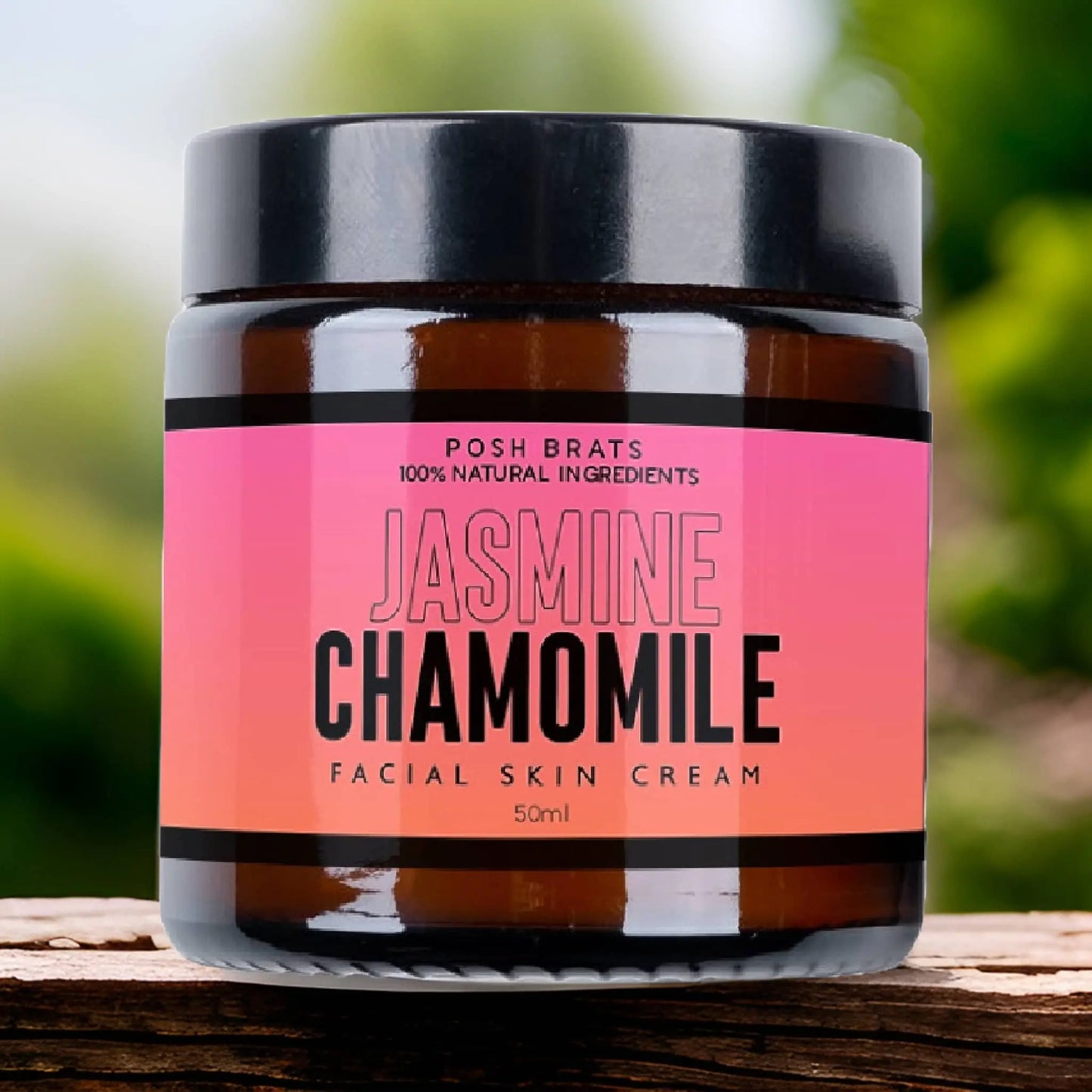 A 50ml jar of Posh Brats’ Jasmine Chamomile Skin Cream, with 100% natural ingredients, provides deep hydration and anti-aging benefits. It features a pink label with black and white text, resting on wood with greenery blurred in the background.