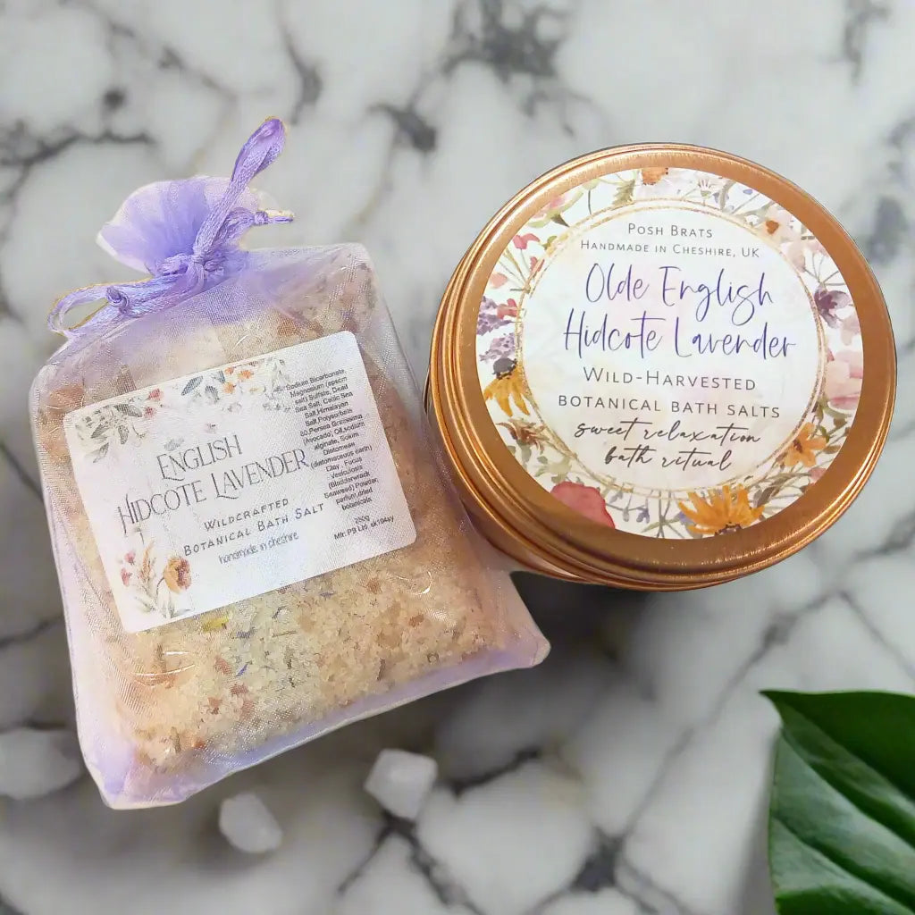 A bag of Posh Brats English Hidcote Lavender Botanical Bath Salt and a matching tin with a copper lid, adorned with floral designs, rest on a marble surface. Eucalyptus leaves add natural tranquility to this relaxation duo.