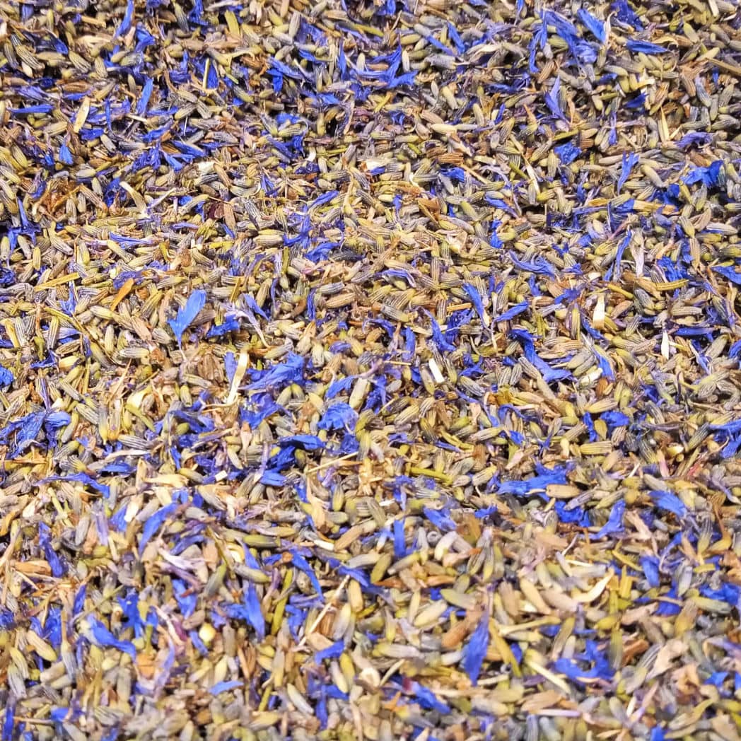 A close-up of Wildcraft Botanical Potpourri by Posh Brats reveals aromatic herb and flower mixtures, including dried lavender buds and small purple petals, all displayed in a blend of muted green and purple hues for a textured, colorful pattern.