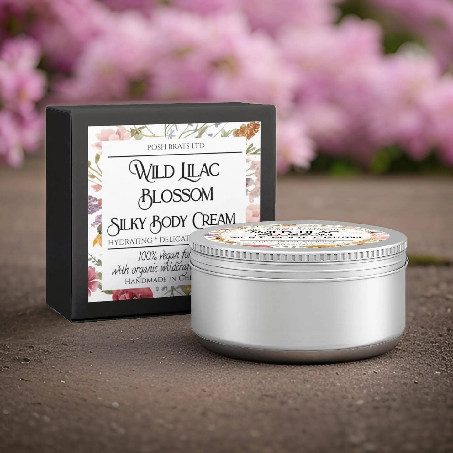A jar of Posh Brats Wild Lilac Blossom Silky Body Butter Cream sits elegantly on stone beside its box, with blurred pink flowers framing it. This luxurious vegan and organic skincare product promises a lavish experience for your skin.