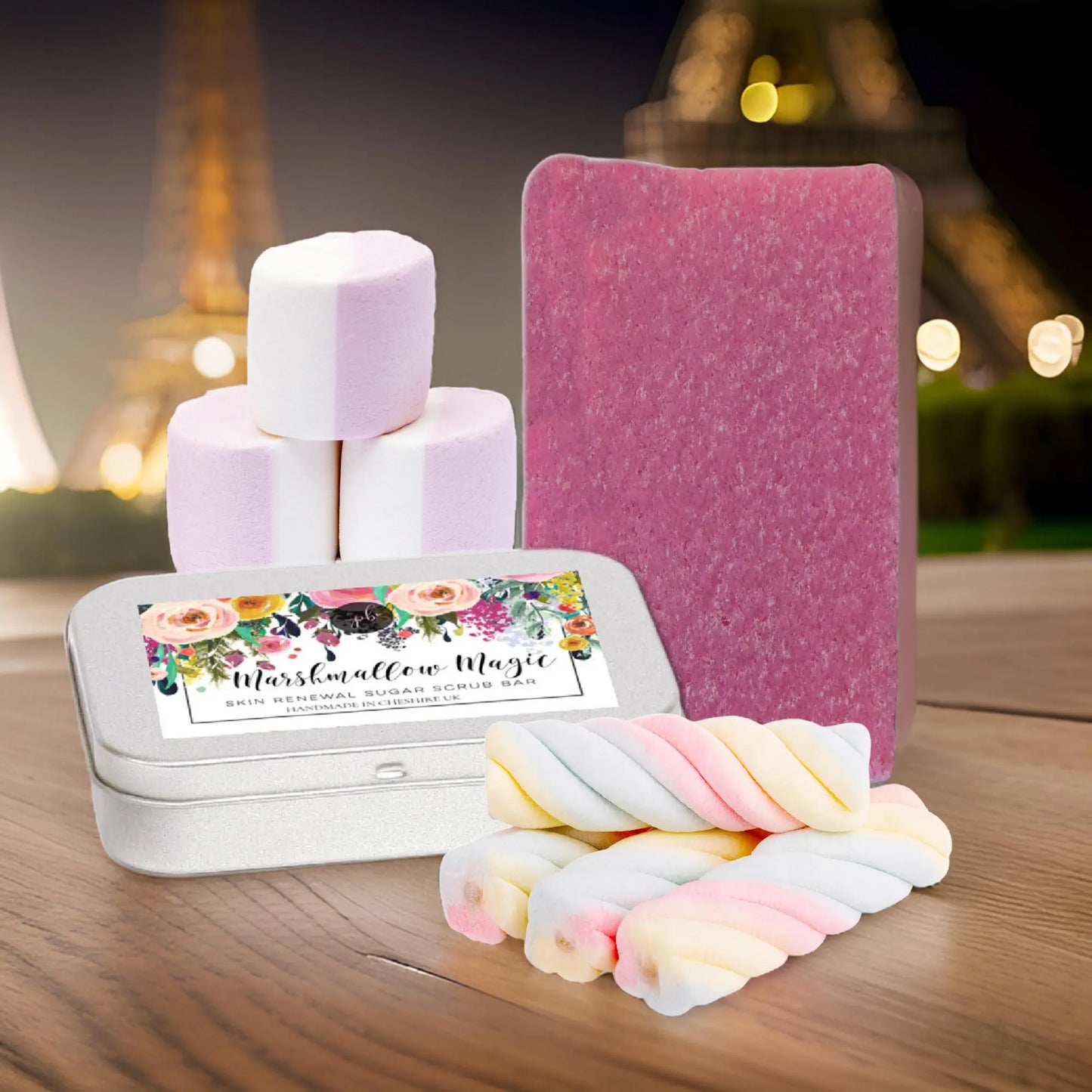 Beside a tin labeled Marshmallow Magic, a stack of pink and white marshmallows, a Marshmallow Magic Sugar Scrub Bar by Posh Brats, and colorful twisted marshmallows enhance the dreamy Eiffel Tower backdrop, promising a sweet exfoliation experience.