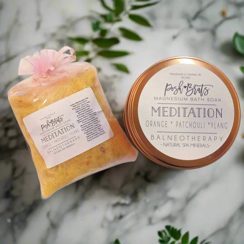 A pouch and tin of Posh Brats Meditation Aromatherapy Bath Salt, infused with Himalayan pink salt and essential oils, rest elegantly on a marble surface. The label showcases scents of orange, patchouli, ylang and emphasizes spa minerals for ultimate relaxation through balneotherapy.