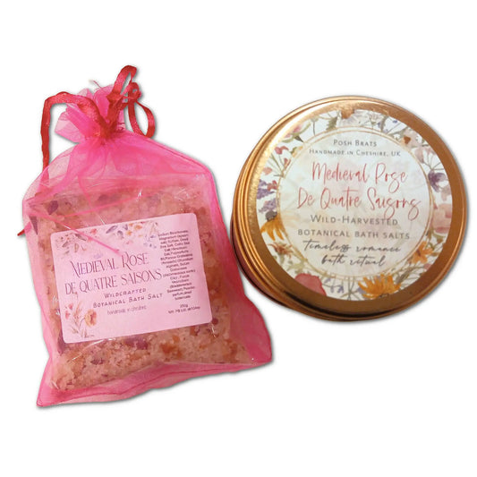 A pink mesh bag of Posh Brats Medieval Rose De Quatre Saisons Botanical Bath Salt, made with an ancient formula, is next to an open tin with a labeled lid displaying similar information. The scene is set against a pristine white background.