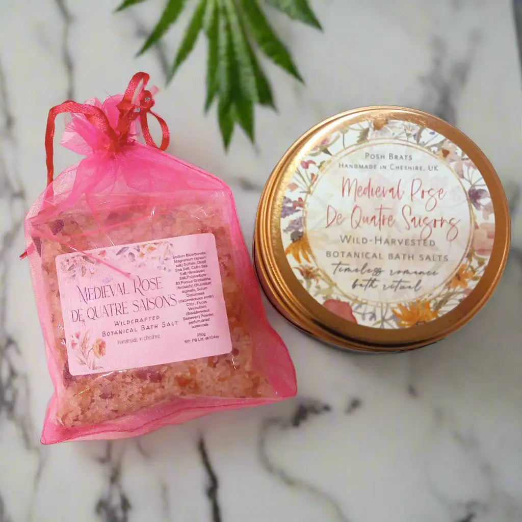 A pink mesh bag of Medieval Rose De Quatre Saisons bath salts, crafted by Posh Brats, lies next to a round tin with a floral design, both embodying an ancient formula. A green plant leaf peeks from the background.
