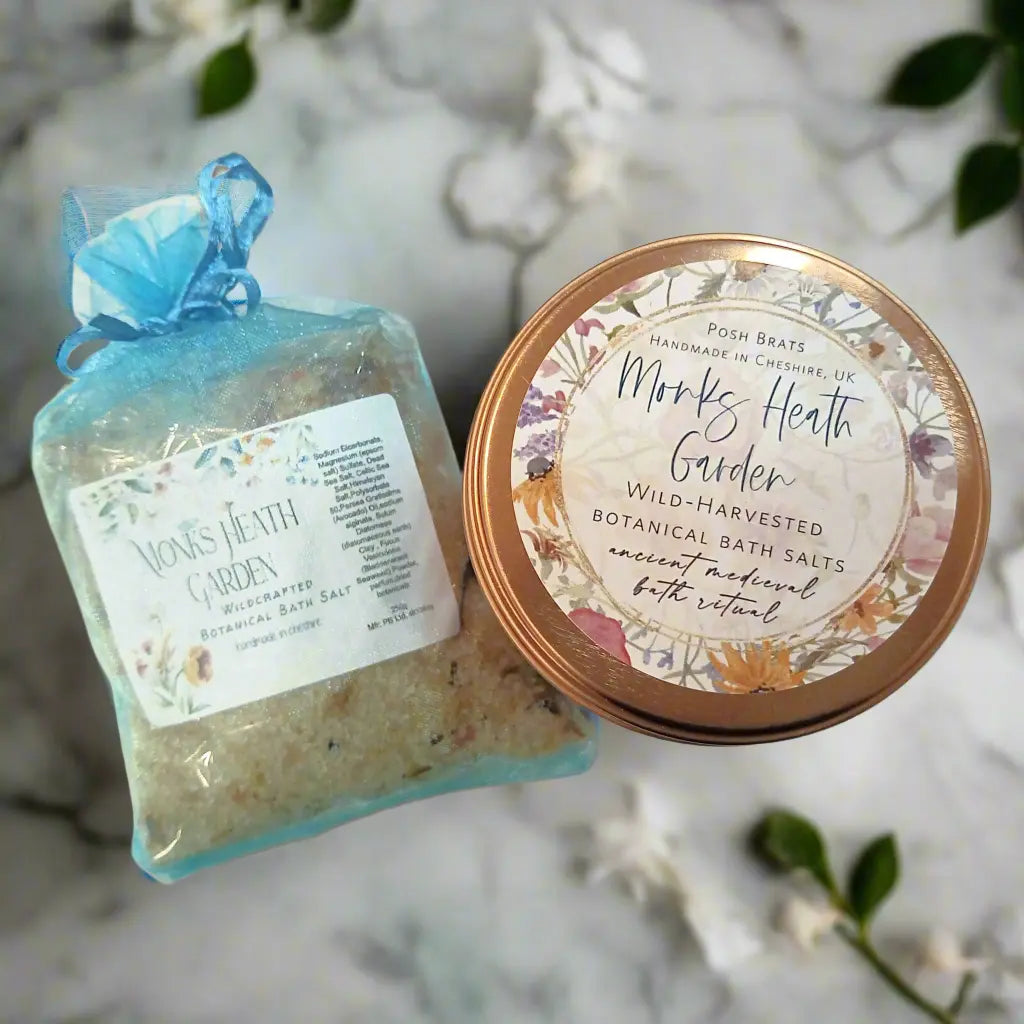 An overhead shot of a Posh Brats Monks Heath Garden bath set includes a Lavender and Rosemary bath salt pouch and a Medieval Botanical Bath Salt tin. The items sit on a marble surface sprinkled with white flowers.