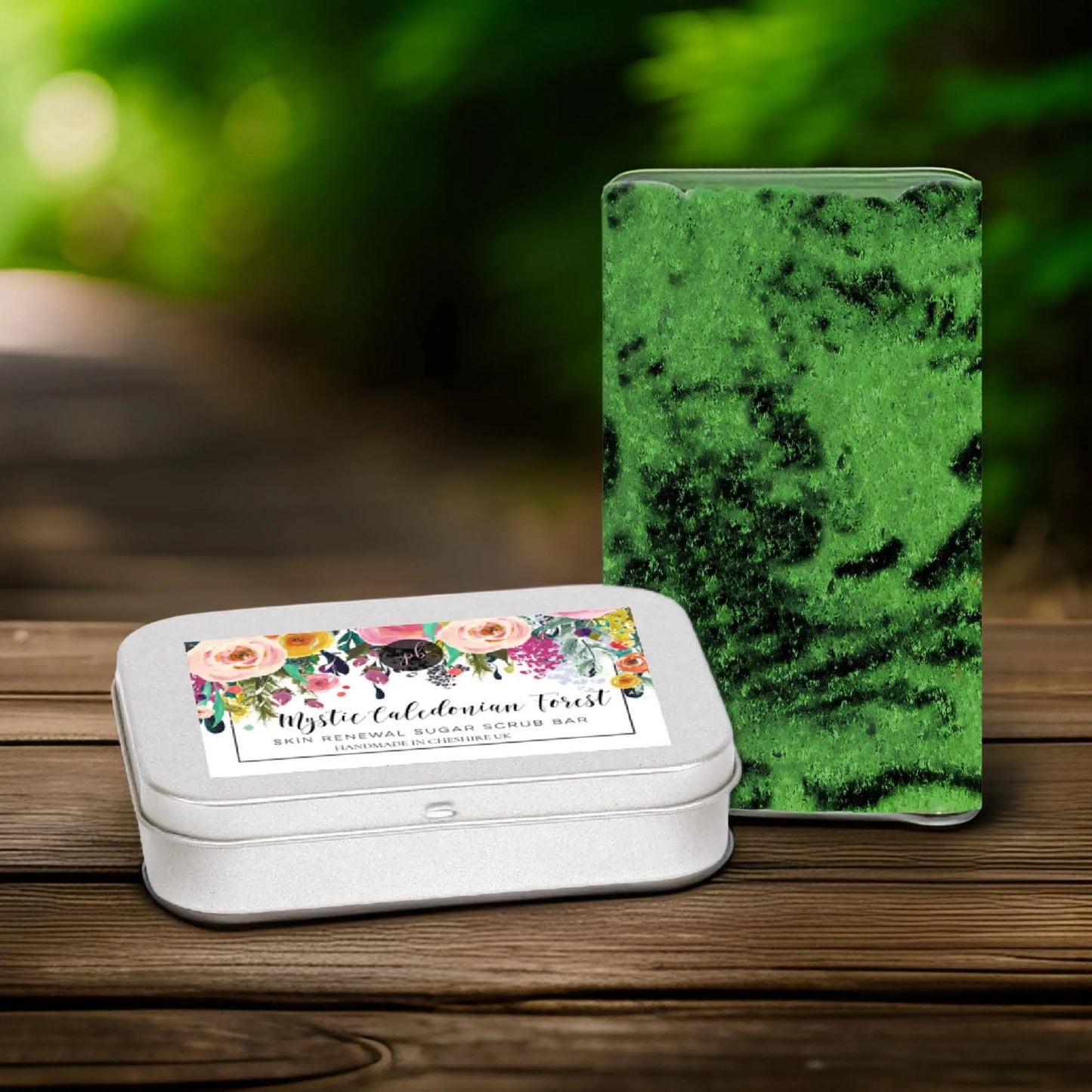 A decorative metal tin and a green, textured Mystic Caledonian Forest Sugar Scrub Bar from Posh Brats rest on a wooden surface against a blurred, leafy backdrop, evoking the essence of Scottish botanicals.