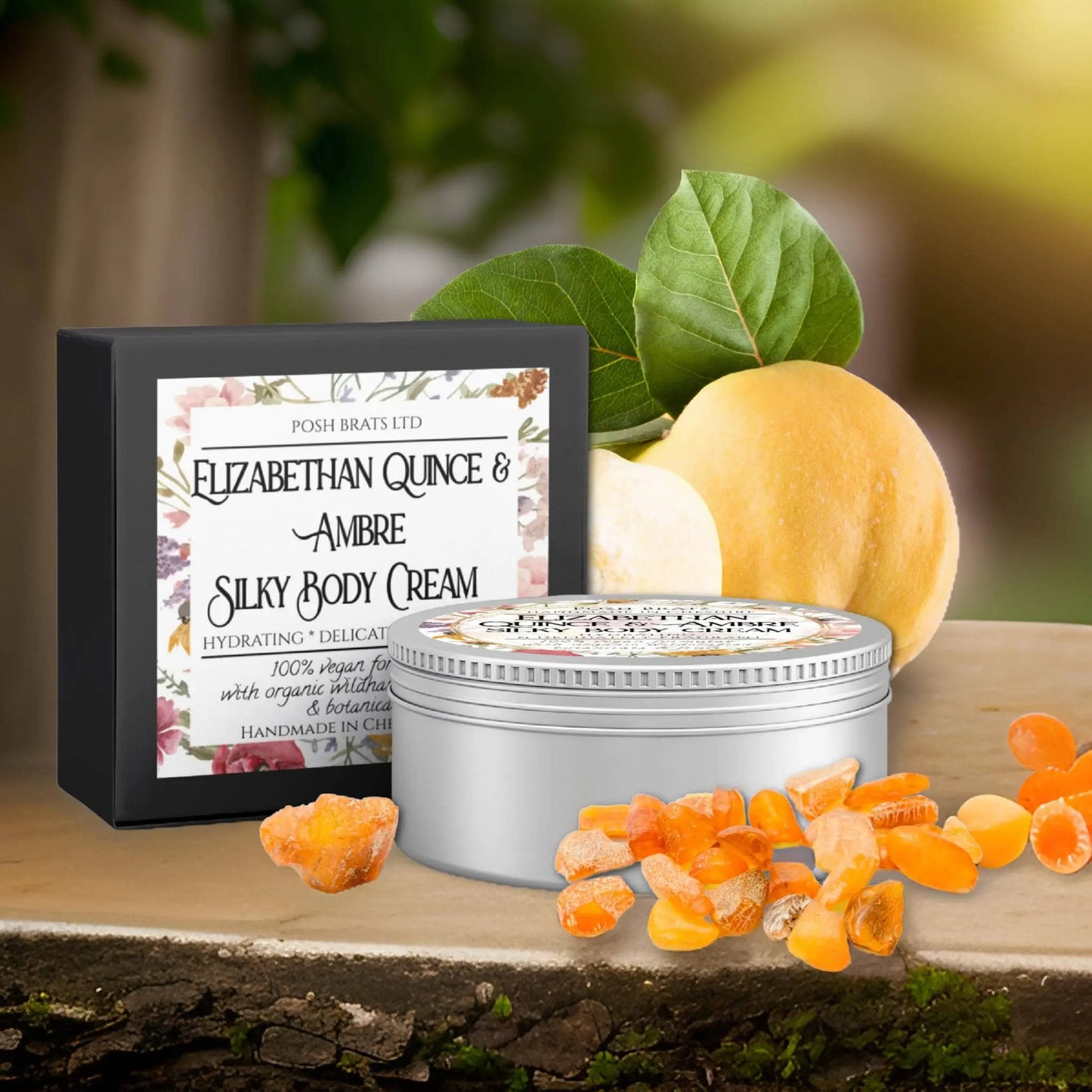 A jar and box of Posh Brats Elizabethan Quince + Ambre Silky Body Butter Cream sit elegantly on stone, accented with quince fruit, Baltic amber stones, and green leaves. The packaging features floral patterns and decorative text.