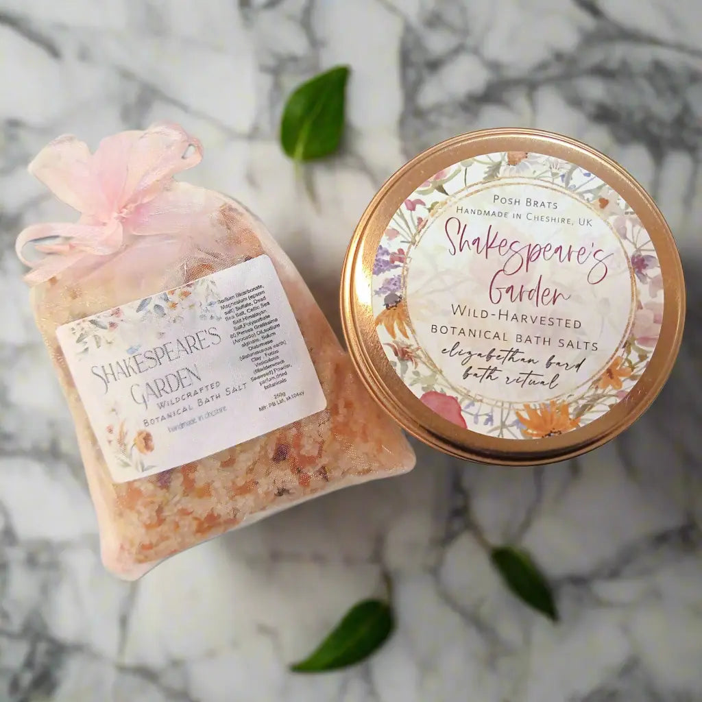 A pouch and a tin of Posh Brats Shakespeares Garden Wildcraft Botanical Bath Salt with floral labels rest elegantly on marble, exuding lavenders calming essence. A green leaf adds tranquility nearby.