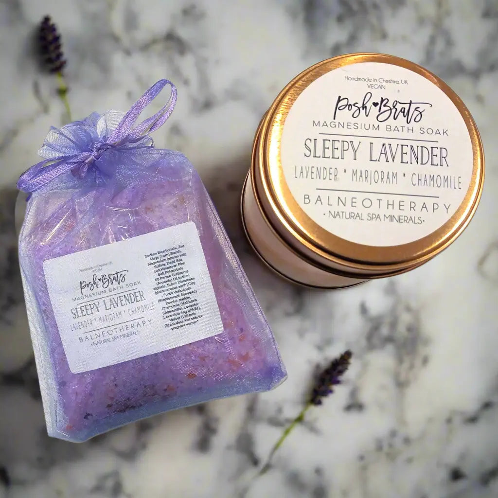 A Posh Brats Sleepy Lavender Botanical Bath Salt jar with a metallic lid, containing lavender essential oil, marjoram, and chamomile, sits on marble next to a semi-transparent sachet with a purple hue.