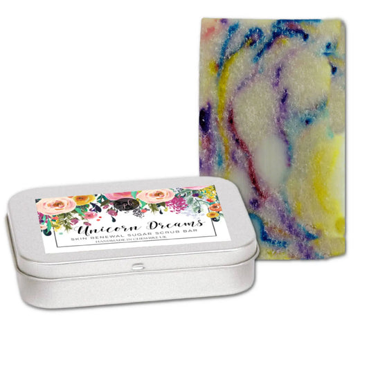 A Posh Brats Unicorn Dreams Sugar Scrub Bar, a vibrant blend with rainbow swirls, is placed beside its floral-designed, vegan skincare tin labeled Unicorn Dreams. It promises gentle exfoliation and vibrant freshness.