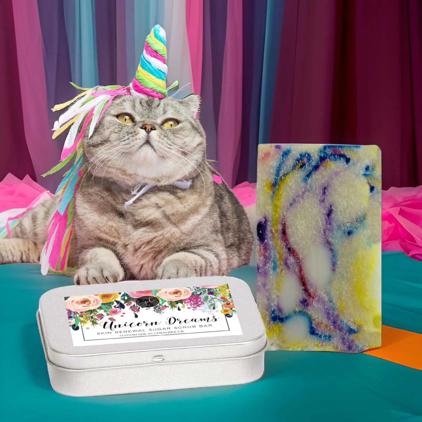 A fluffy gray cat with a colorful unicorn horn hat sits beside a decorative tin labeled Unicorn Dreams, featuring Posh Brats vibrant Unicorn Dreams Sugar Scrub Bar for vegan skincare, all set against fabric in various hues.