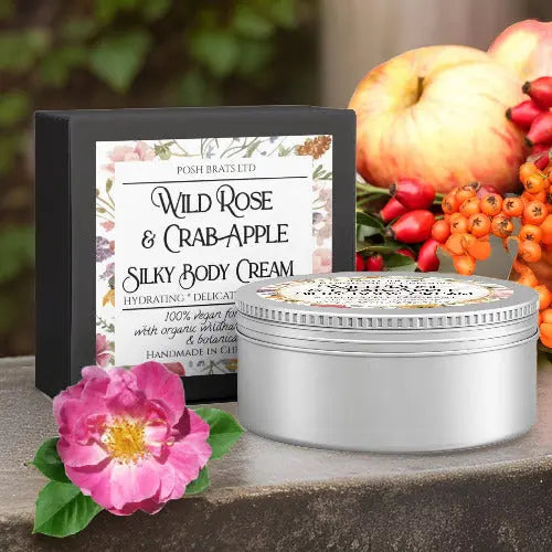 A tin of Wild Rose & Crab Apple Silky Body Butter Cream by Posh Brats is displayed with its decorative box. The background, featuring roses, apples, and orange berries on a stone surface, creates a natural, rustic vibe.