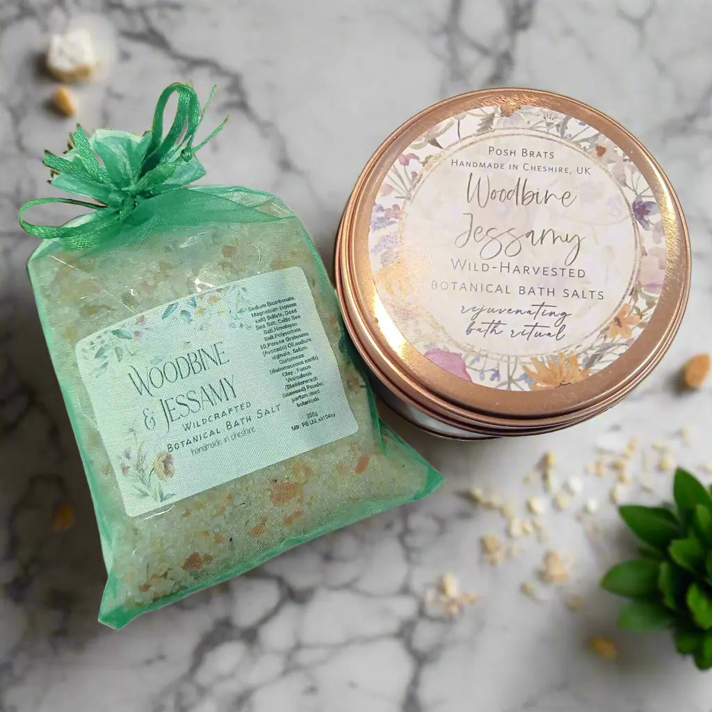 A tin labeled Wild-Harvested and a transparent, green-accented pouch of Posh Brats Woodbine Jessamy Botanical Bath Salt sit on a marbled surface, offering relaxation with their floral design and enticing contents.