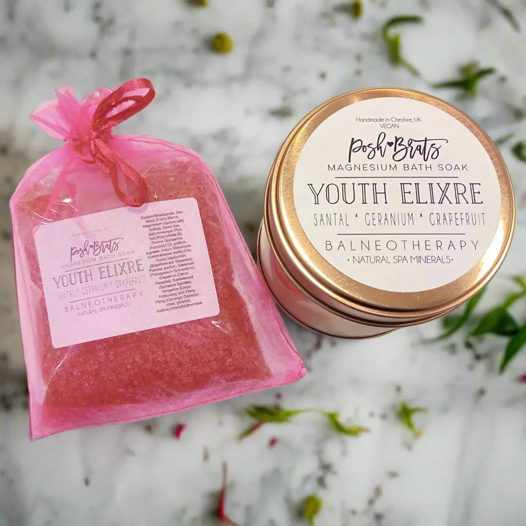 A pink organza bag with Youth Elixre Magnesium Aromatherapy Bath Salt by Posh Brats sits next to a matching lidded jar on marble. Infused with essential oils, it offers santal, geranium, and grapefruit scents plus spa minerals and Dead Sea salt for a serene experience.