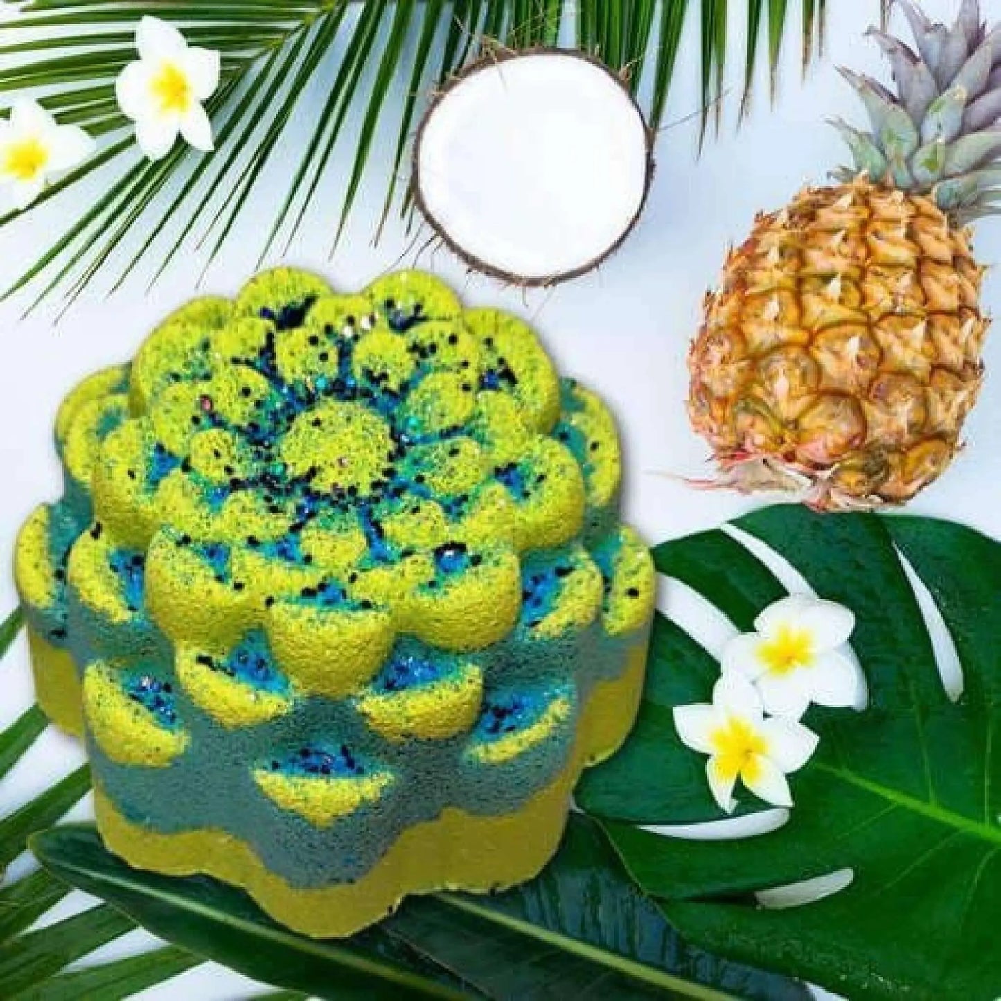 A green and blue flower-shaped cake surrounded by pineapple, coconut halves, tropical leaves, and white plumeria blooms evokes an Aloha Paradise Fizzy Bath Bomb-inspired getaway.