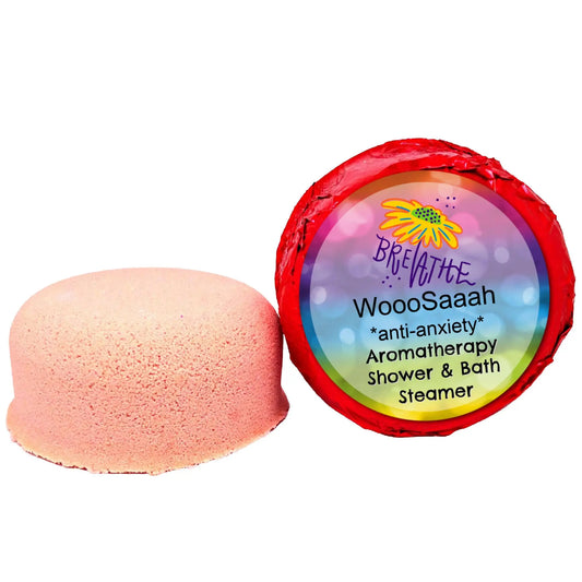 A pink, round shower steamer infused with essential oils is unwrapped beside its vibrant red and rainbow packaging labeled Breathe, from the WooSaah Anti-Anxiety Aromatherapy line by Posh Brats.