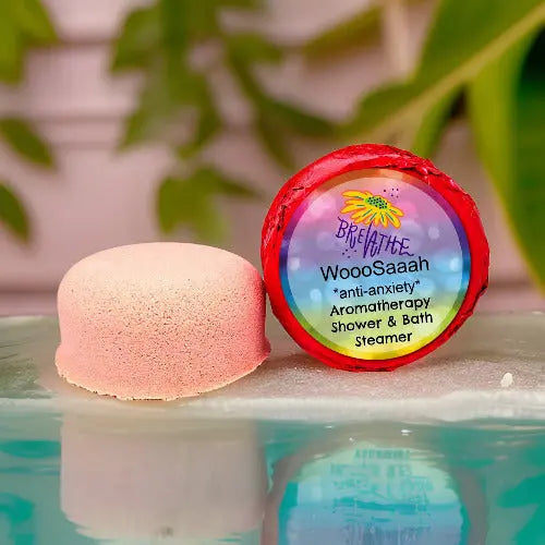 A pink WooSaah Anti-Anxiety Aromatherapy Shower Steamer from Posh Brats rests on a reflective surface. The packaging features its essential oils blend for anti-anxiety relief, set against a backdrop of soothing blurred green leaves.
