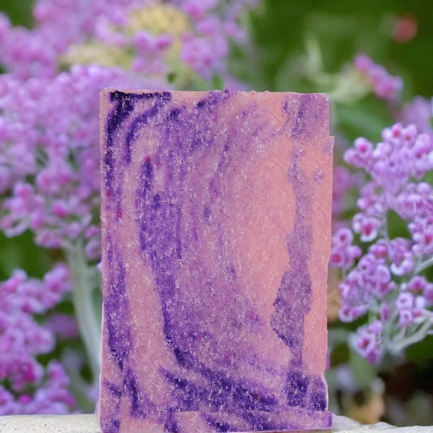 The Berry Comforting Sugar Scrub Bar by Posh Brats features a pink base with purple swirls, standing among pink and purple flowers with lush greenery. It provides gentle exfoliation with vegan-friendly care.