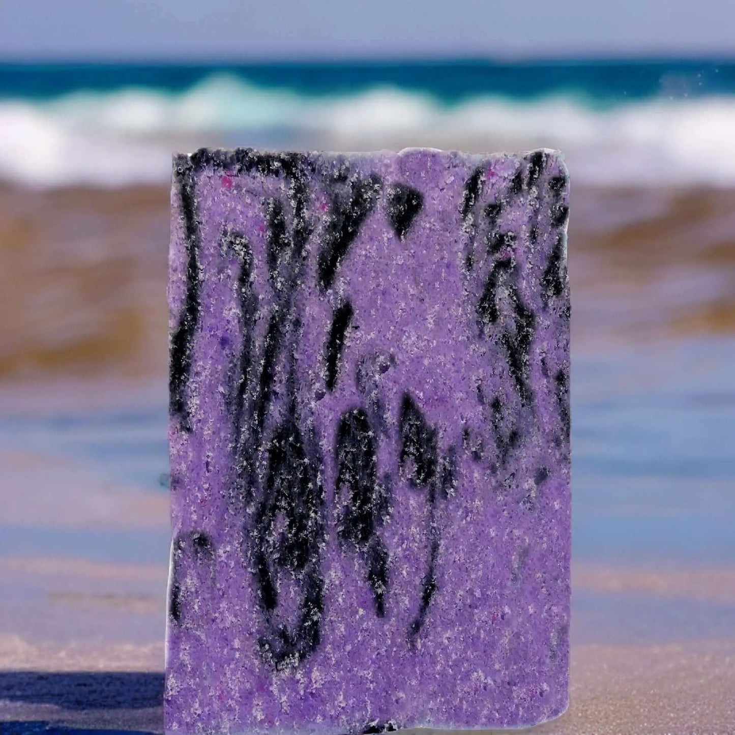 A rectangular bar of purple and black marbled soap, resembling the Posh Brats Black Pearl Sugar Scrub Bar, stands upright on a sandy beach with waves in the background.