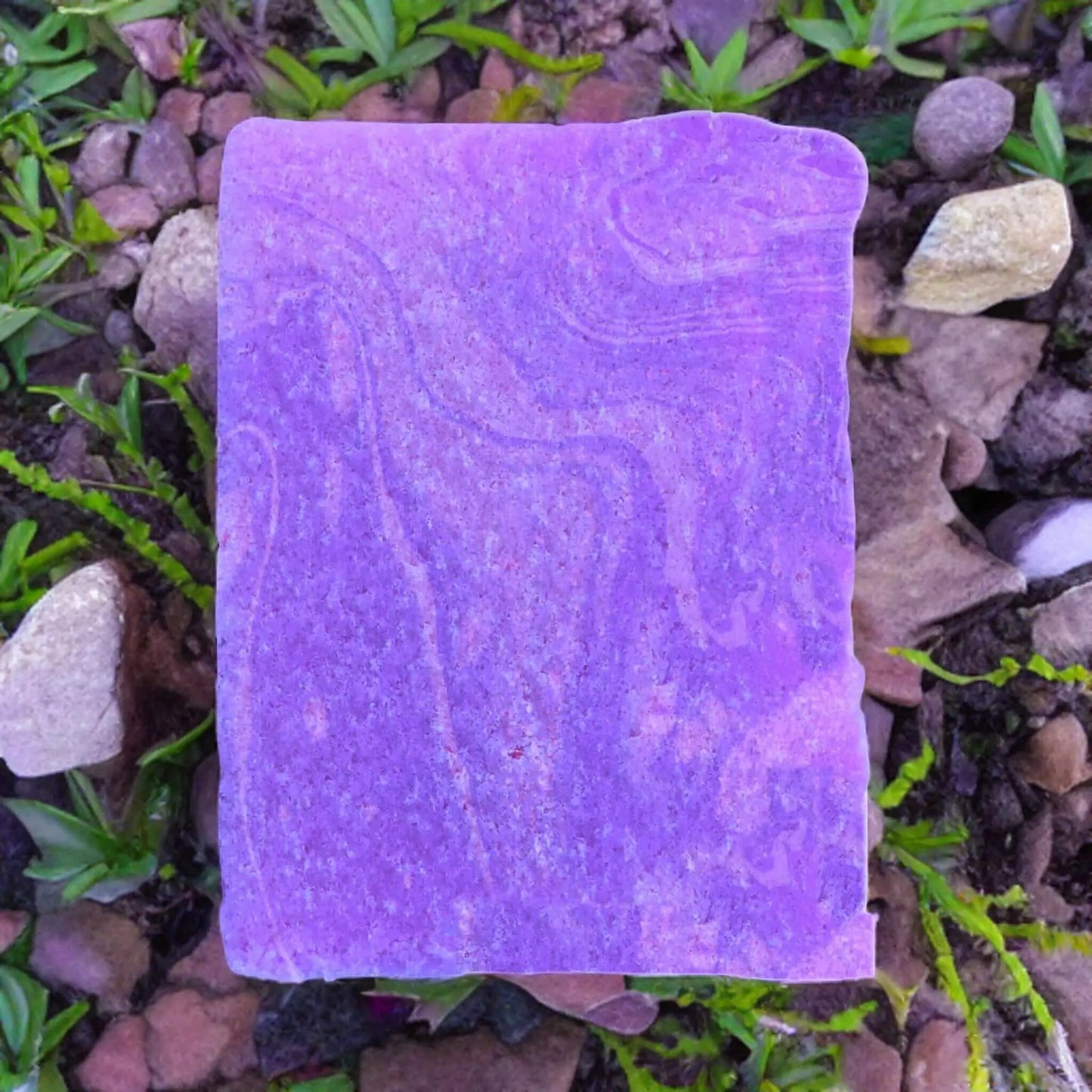 A purple rock with swirling patterns, surrounded by small rocks and green plants, evokes the soft hues of Posh Brats Celtic Sea Lavender Sugar Scrub Bar.