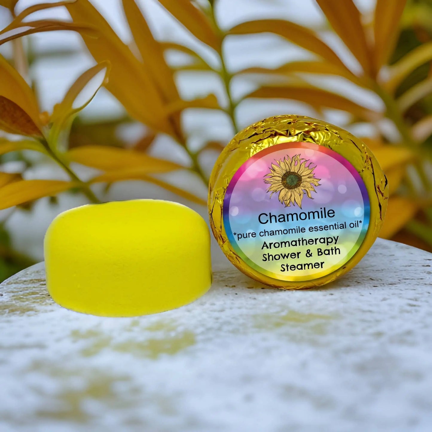 The Posh Brats Chamomile Aromatherapy Shower Steamer, featuring German Blue oil and pure chamomile essential oil, sits on a marble surface with yellow-green foliage in the background. Its label is decorated with a sunflower.