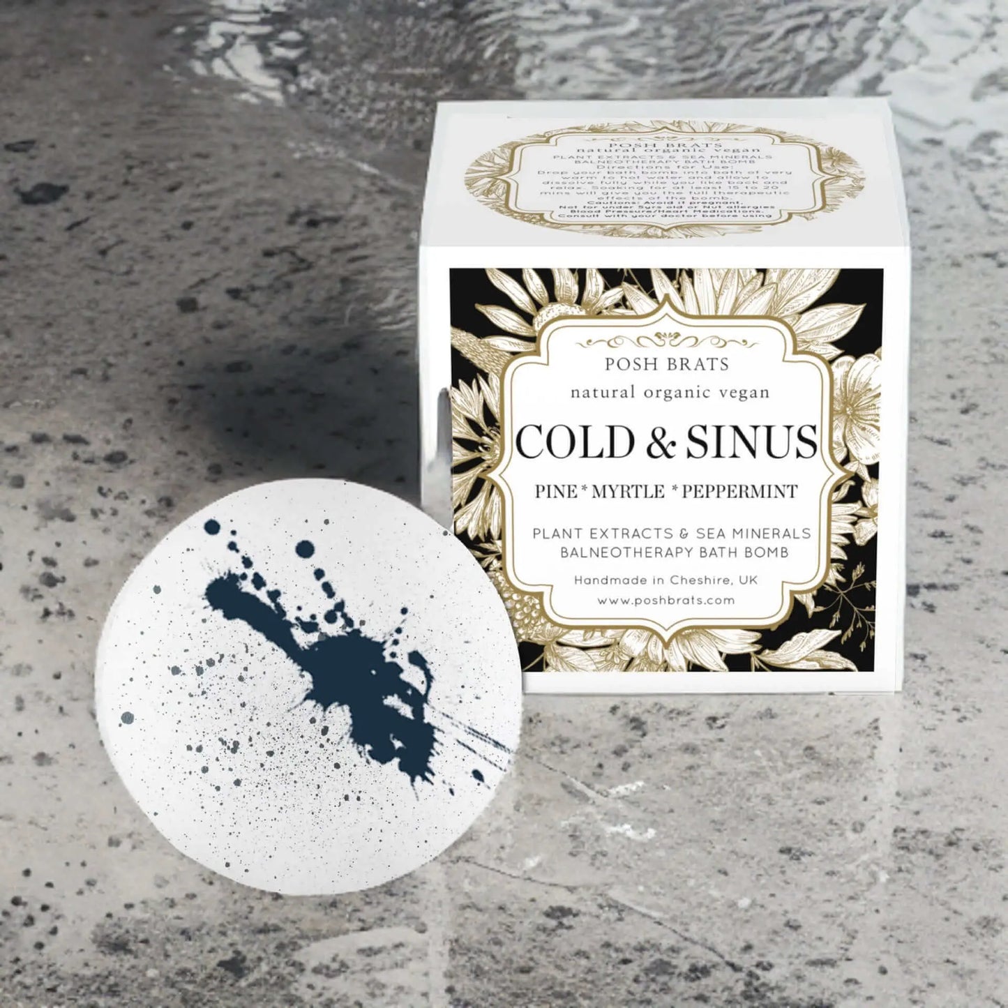 A Cold Sinus Aromatherapy Bath Bomb by Posh Brats, featuring black splatter patterns, rests beside its box. Made with natural, organic, vegan ingredients like pine, myrtle, and peppermint essential oils, it offers sinus congestion relief.