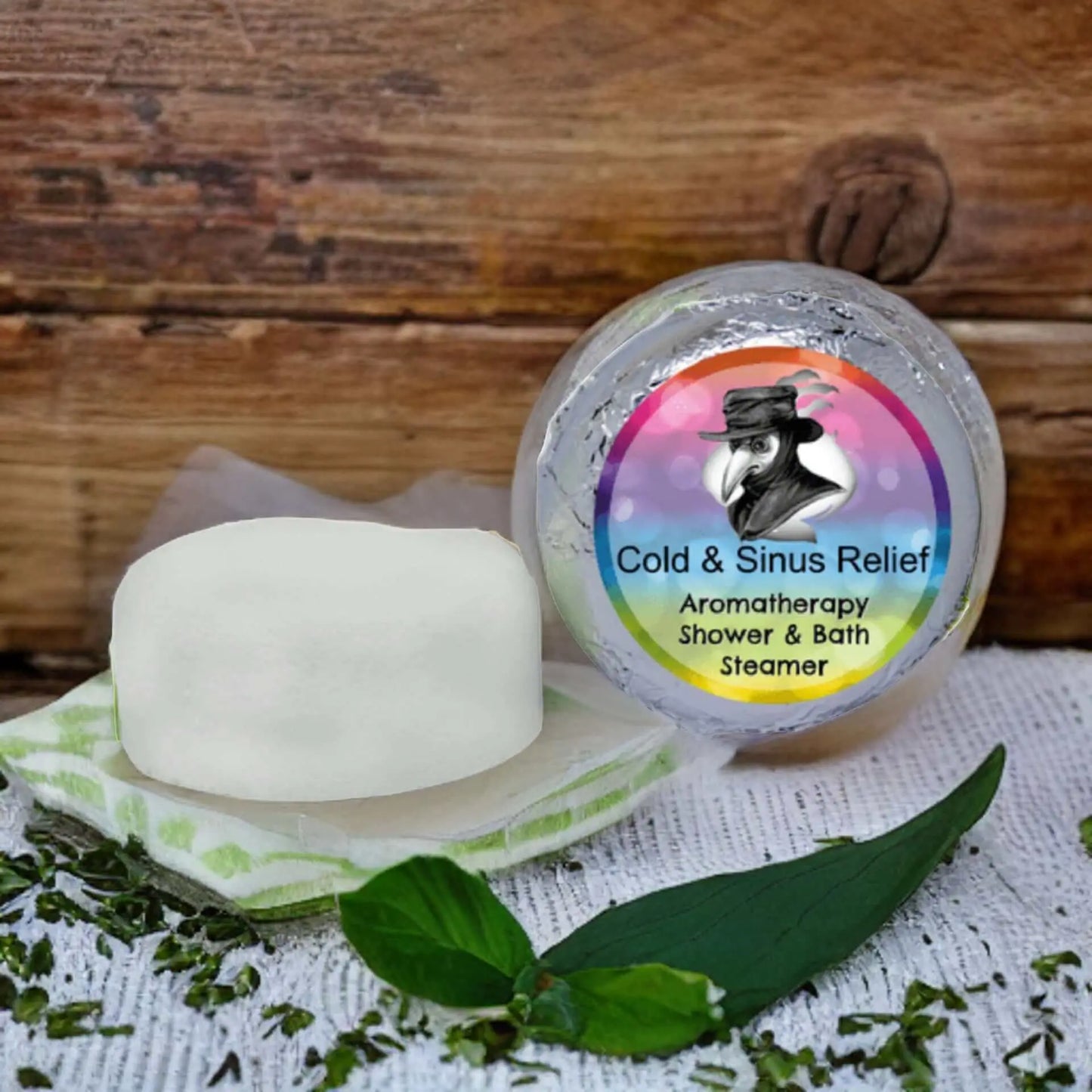 A round container of Posh Brats Cold Sinus Aromatherapy Shower Steamer sits on wood, promising congestion relief. Nearby, a white soap bar rests with fresh green leaves and herbs, enhancing natural tranquility.
