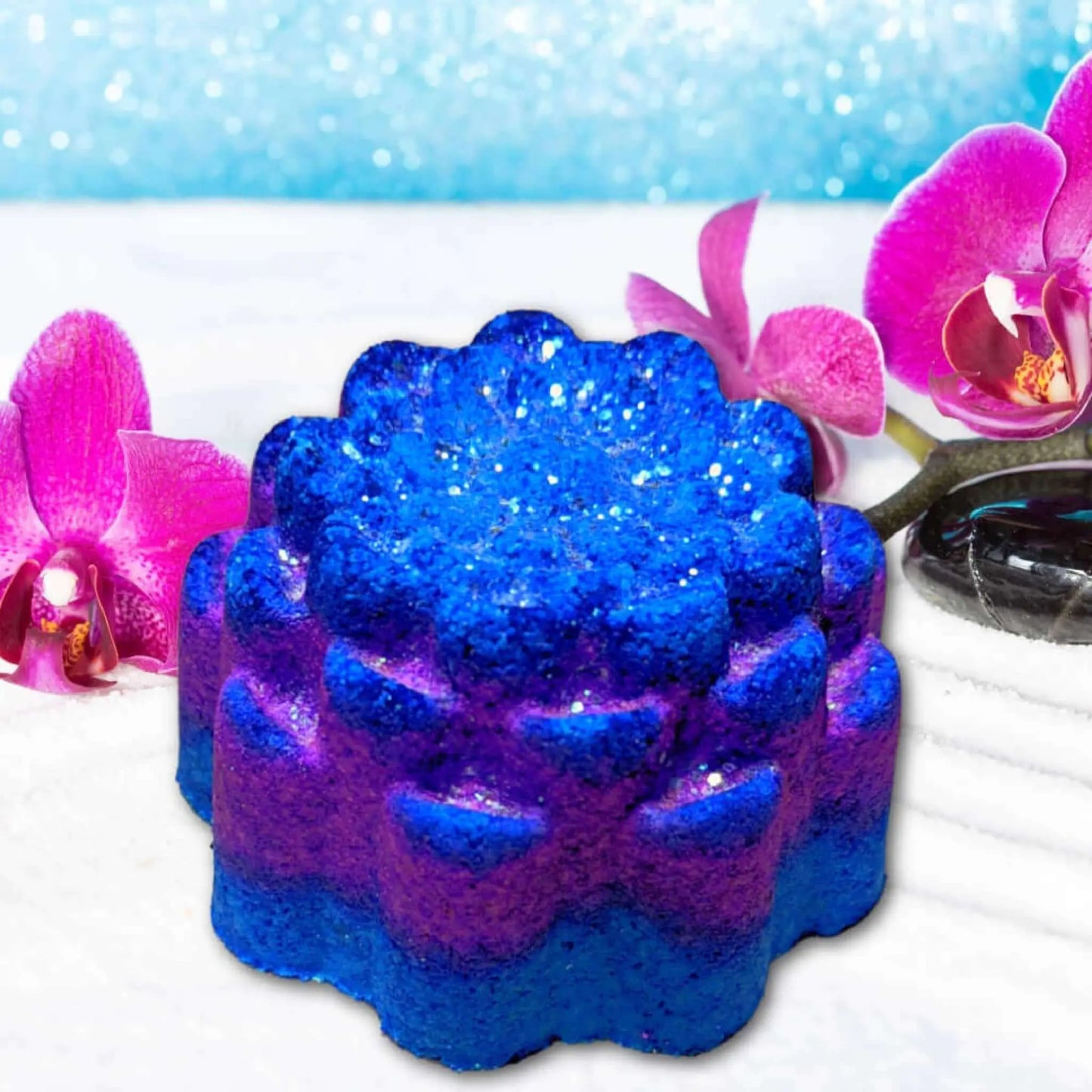 The Desert Spa Fizzy Bath Bomb by Posh Brats, a vibrant blue and purple floral-shaped delight, rests on a white surface with pink orchids nearby. The background shimmers with blue sparkles, promising a rejuvenating soak with this luxury bath accessory.