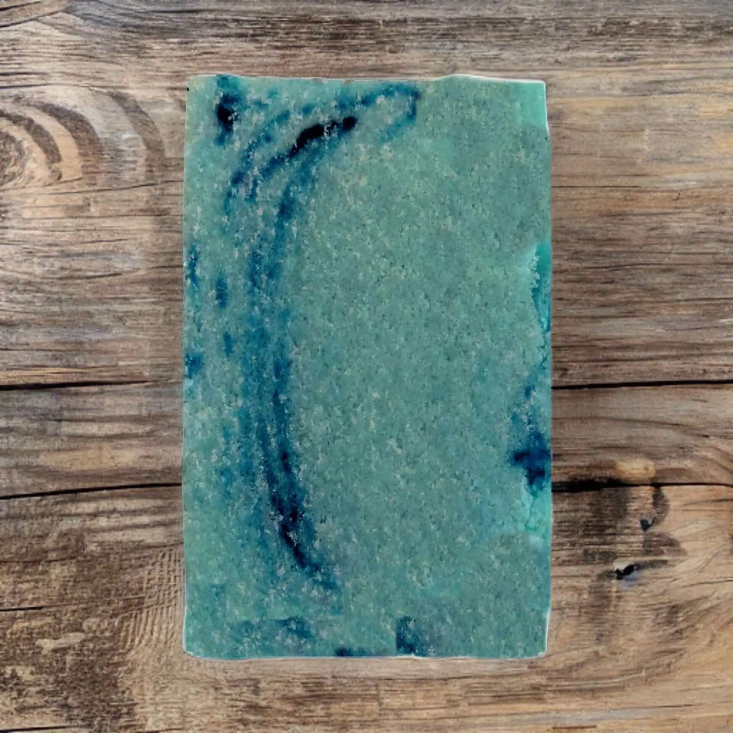 The Dirty Hippy Patchouli Sugar Scrub Bar by Posh Brats, a square green soap with dark blue swirls, rests on a rustic wooden surface.