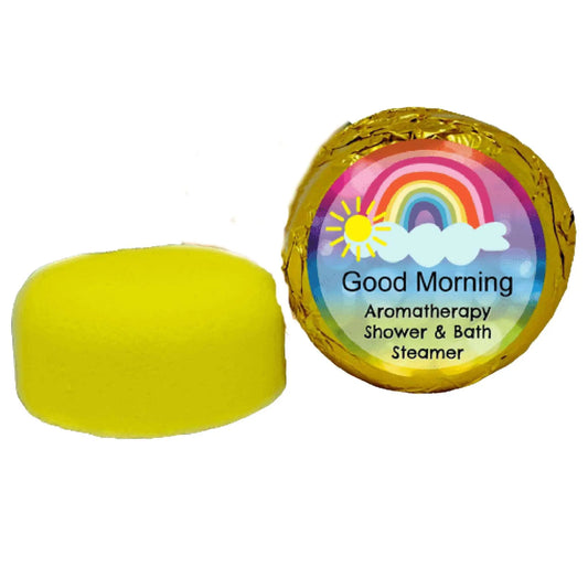 A yellow essential oils shower steamer by Posh Brats sits beside its packaging, wrapped in gold foil with a vibrant sun, cloud, and rainbow label. The text reads: Good Morning Aromatherapy Shower Steamer.